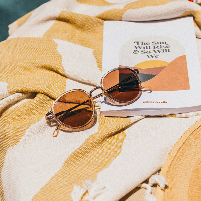 Sunglasses on a book titled 'The Sun Will Rise & So Will We' on a yellow and white patterned fabric.