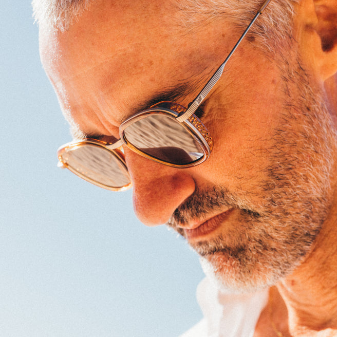 Bernina - Desert Amber - Close-up of a person wearing gold-rimmed sunglasses against a clear blue sky.