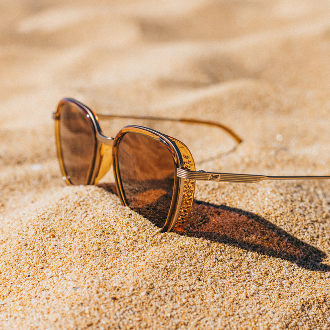 Bernina - Desert Amber - Gold-framed sunglasses on a sandy surface