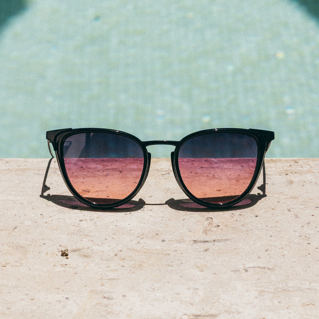 Sunglasses with pink and purple gradient lenses on a wooden surface with a pool in the background