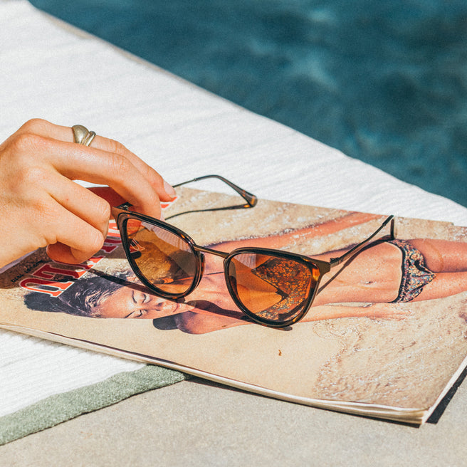 Person holding sunglasses over a magazine by a pool