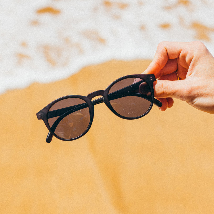 Person holding sunglasses over sand with ocean waves in the background