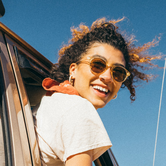 Woman with sunglasses smiling while leaning out of a car window against a blue sky.