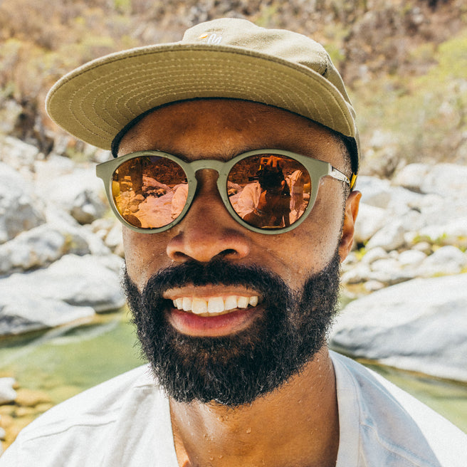 Man wearing sunglasses and a cap with a scenic reflection, standing by a river.