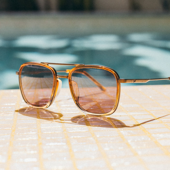 Gold-framed sunglasses on a woven surface with a blurred pool background