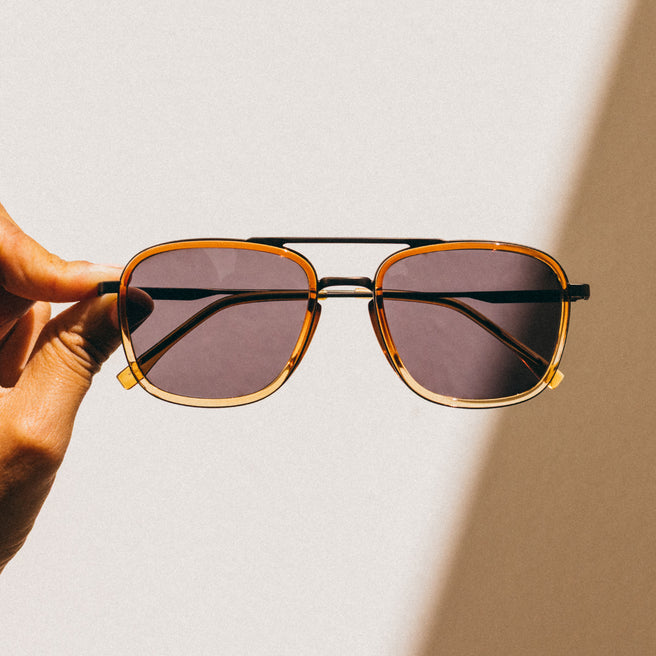 Sunglasses with gold frame and dark lenses held by a hand against a light background
