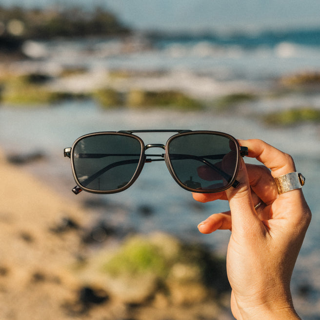 Estero - Olive Forest - Person holding sunglasses with a blurred beach background