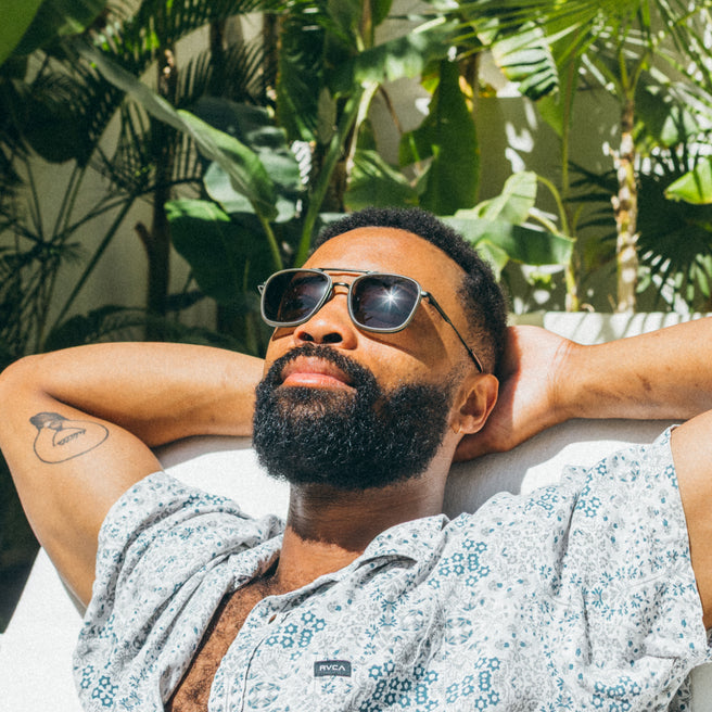Estero - Olive Forest - Man wearing sunglasses and a patterned shirt, lying on a couch with plants in the background