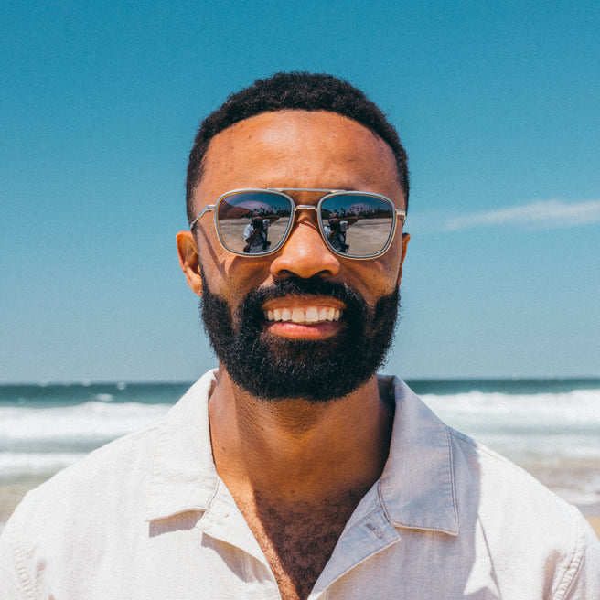 Estero - Storm Honey Silver - Man wearing sunglasses and a light shirt on a beach with clear blue sky and ocean.