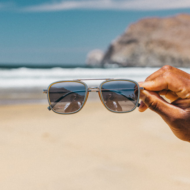 Estero - Storm Honey Silver - Sunglasses held by a hand with a beach and ocean in the background