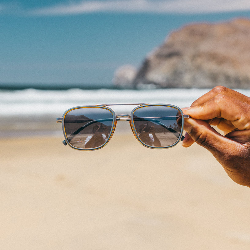 Sunglasses held by a hand with a beach and ocean in the background