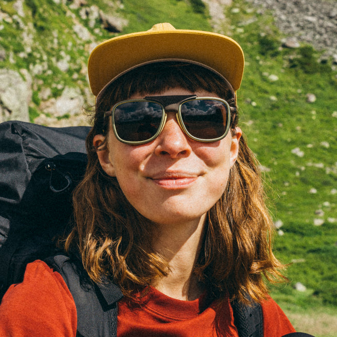 Person wearing sunglasses and a yellow cap with a backpack, standing in a mountainous area.