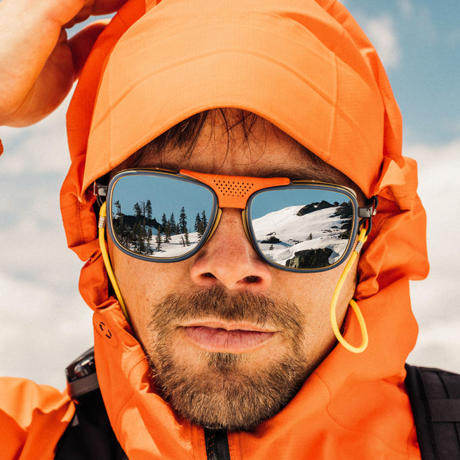 Person wearing an orange hooded jacket and sunglasses with a mountain reflection, standing against a snowy landscape.
