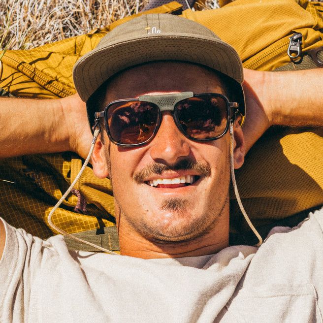 Man wearing sunglasses and a cap with a yellow backpack, lying on the ground.