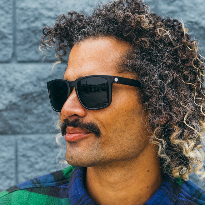 Man wearing sunglasses with a plaid shirt against a gray wall