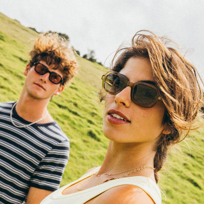 Two people wearing sunglasses with a grassy hill in the background
