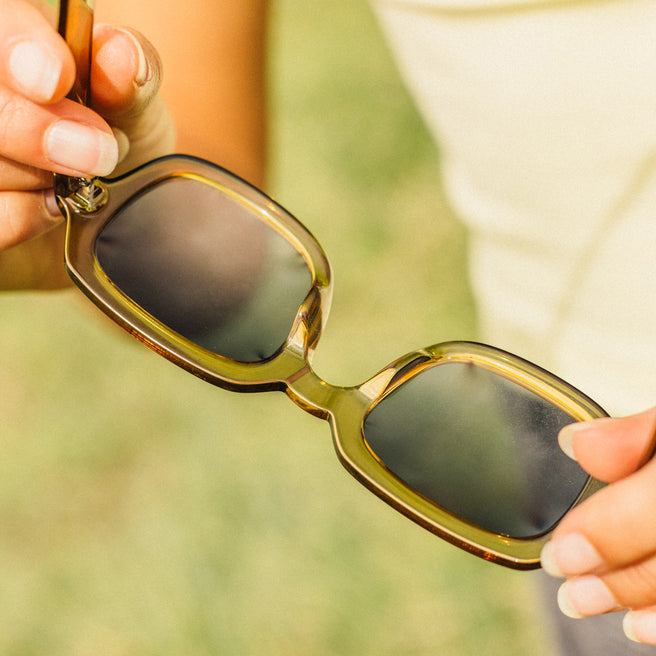Gold-framed sunglasses held by a person with a blurred natural background