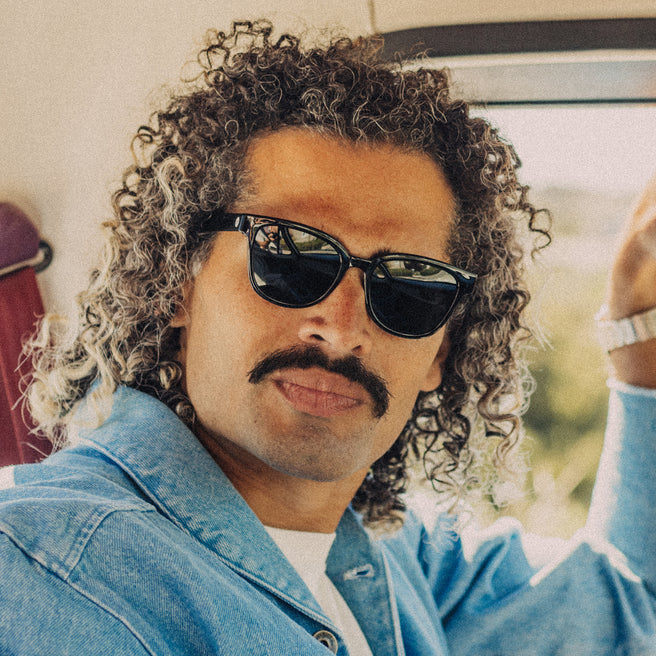 Man with curly hair wearing sunglasses and a blue jacket inside a vehicle.