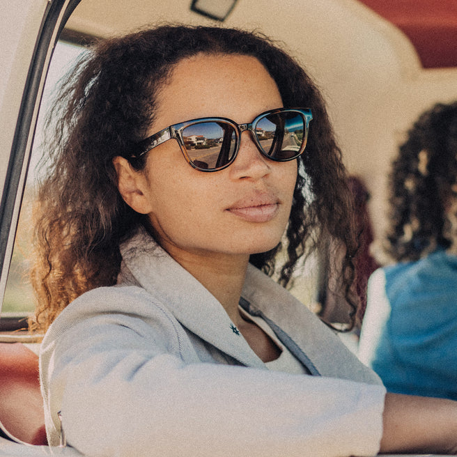 Woman wearing sunglasses sitting inside a vehicle with another person in the background.