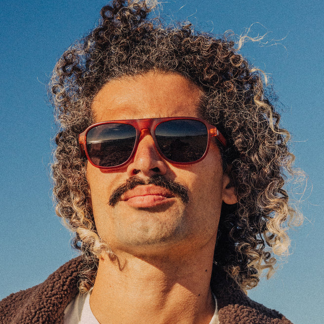 Man with curly hair and mustache wearing red sunglasses against a blue sky.