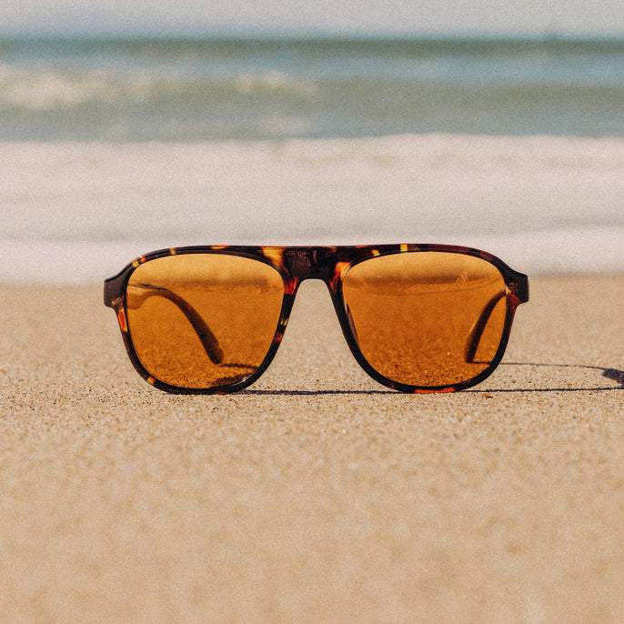 Sunglasses with orange lenses and tortoiseshell frame on a sandy beach.