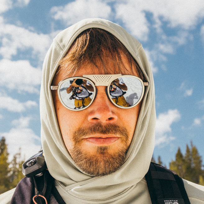Person wearing a hooded jacket with reflective sunglasses, standing against a blue sky with clouds and trees.
