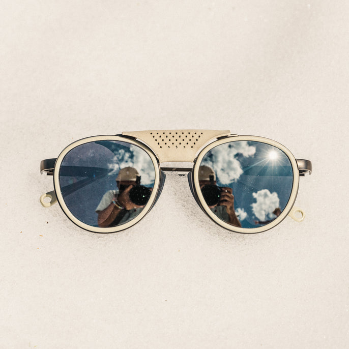 Sunglasses with reflective lenses showing a blue sky and clouds on a beige background