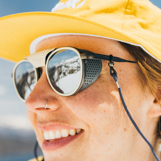 Person wearing a yellow hat and round sunglasses with a mountain reflection.