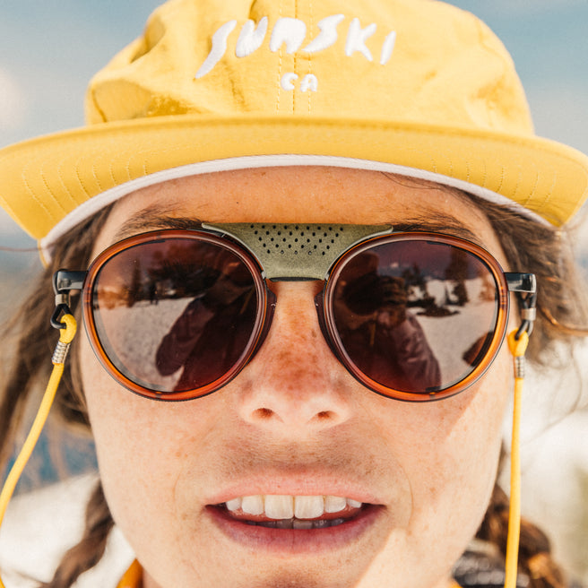 Strada - Rust Amber - Person wearing a yellow 'Sunski' cap and round sunglasses with a blurred background
