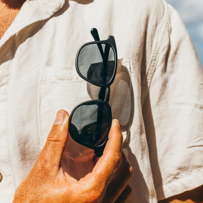 Hand holding a pair of black sunglasses against a light-colored shirt.