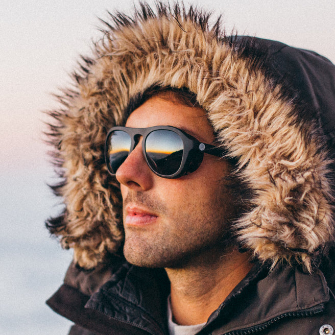 Person wearing a fur-lined hooded jacket and sunglasses against a snowy background
