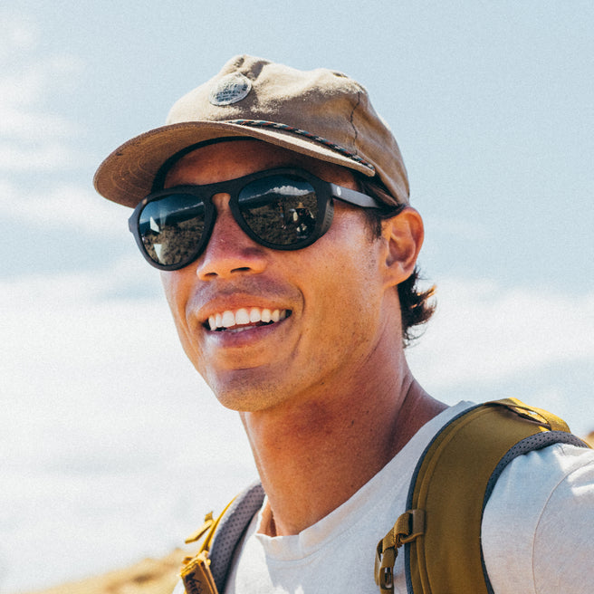 Person wearing sunglasses and a cap with a backpack against a sky background