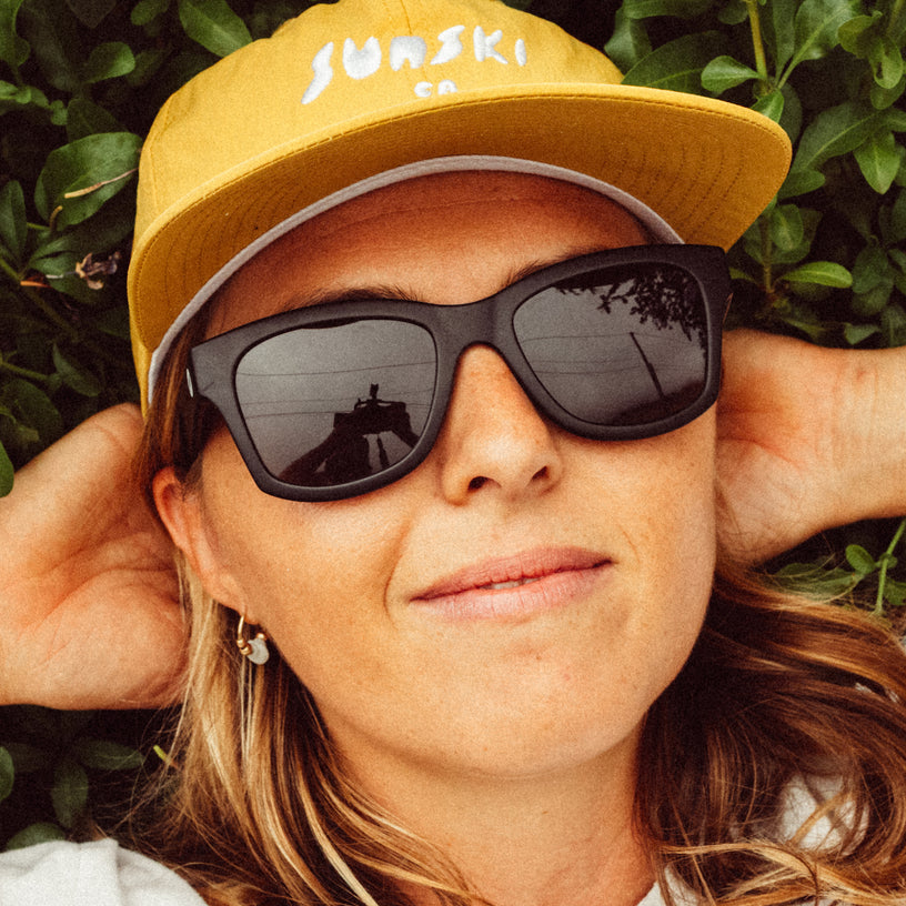 Person wearing a yellow cap with 'Janski' branding and dark sunglasses, lying on green foliage.