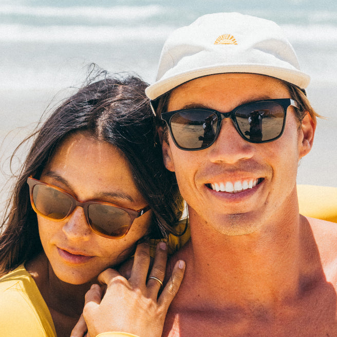 Two people wearing sunglasses and a white cap, standing close together with a beach background.
