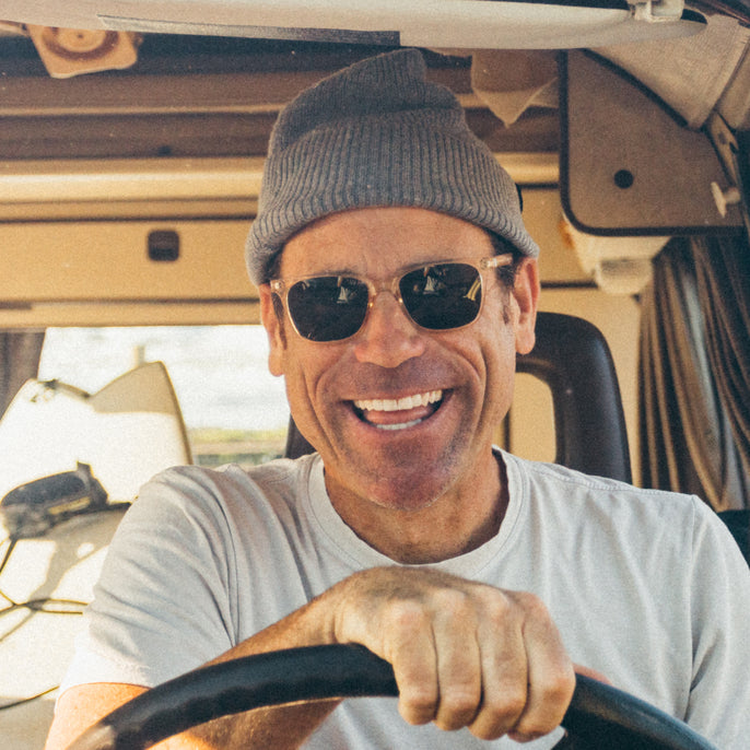 Man driving a vehicle wearing sunglasses and a beanie, smiling.
