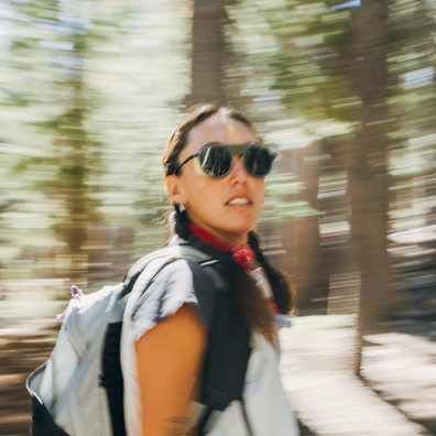A woman wearing black sunglasses, a red neck scarf, and a white backpack walks through a blurred forest path.