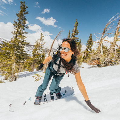 A woman snowboarding on a snowy mountain slope under a blue sky with trees in the background. She is wearing a gray t-shirt, teal pants, a black harness, sunglasses, and a brown glove, with a snowboard visible.