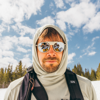 A man wearing a light beige hooded technical jacket and reflective aviator sunglasses, photographed outdoors against a blue sky with clouds and a line of pine trees. A small black label on the chest area reads The North Face.