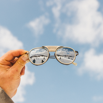 A hand holds a pair of round-frame sunglasses against a blue sky with clouds. The mirrored lenses reflect a snowy outdoor scene.