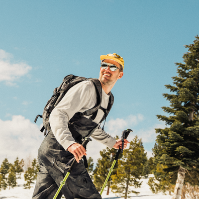 A man hiking outdoors in a snowy landscape under a blue sky, wearing a yellow beanie, sunglasses, a light gray long-sleeve shirt, black bib overalls, and a backpack, while holding ski poles.