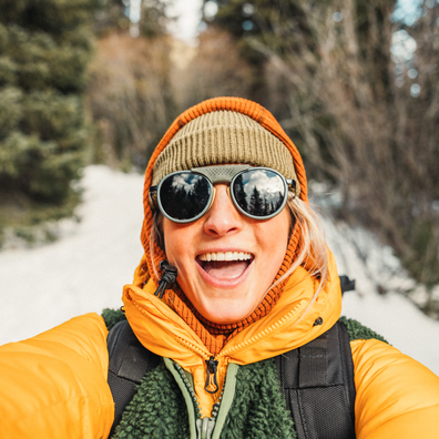 A smiling woman in a snowy outdoor setting takes a selfie while wearing a mustard yellow puffer jacket, a green fleece layer, a tan knit beanie, and round sunglasses.