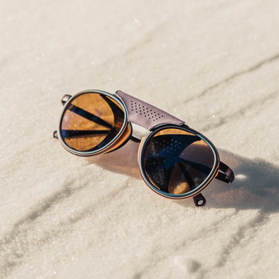 Oversized round sunglasses with reflective amber-tinted lenses and dark metal rims, resting on a sandy beach.