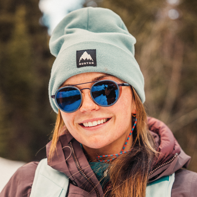 A smiling woman outdoors wearing a light sage green Burton beanie, round blue mirrored sunglasses, and a brown winter jacket. The Burton logo patch is visible on the front of the beanie.