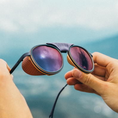 A pair of round sunglasses with amber-brown wood-look frames, black temples, and purple mirrored lenses, held by two hands against a blurred blue sky and mountain background.