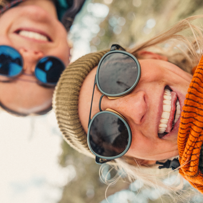 Close-up outdoor selfie of two smiling women wearing round sunglasses. One woman wears a beige knit beanie and the other wears a dark beanie, both with dark-tinted lenses and different frame colors, against a blurred natural background.