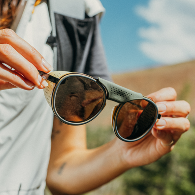 A close-up lifestyle shot of a person outdoors holding a pair of sunglasses with olive green arms and dark brown lenses.