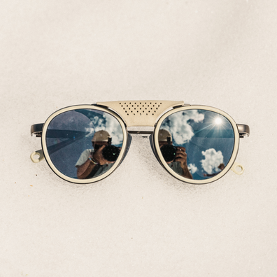 Round sunglasses with a beige perforated metal bridge and silver-toned rims, shown on a textured off-white background. The reflective lenses show a reflection of a person holding a camera.