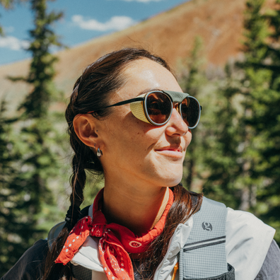 A woman outdoors wearing round sunglasses with a gold-toned frame and dark lenses, a red bandana around her neck, and a gray backpack, with trees and a mountain in the background.