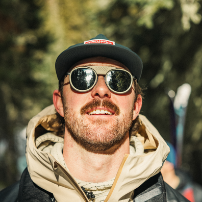 A close-up portrait of a smiling man outdoors wearing round sunglasses with a beige mesh front, a dark green cap with a red logo patch, and a beige hooded jacket. The background is a blurred natural outdoor setting with green foliage.