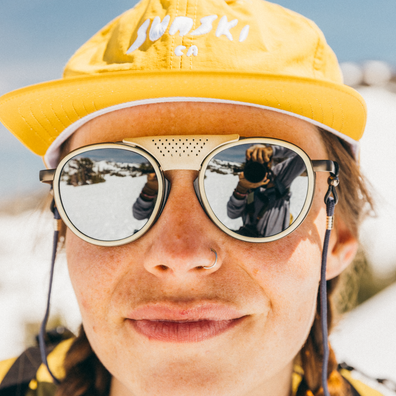 A close-up lifestyle shot of a person wearing round sunglasses with a built-in speaker grille on the bridge, a yellow cap with 'SUNSKI CA' embroidered on it, and a nose ring, with snow visible in the sunglasses' reflective lenses.